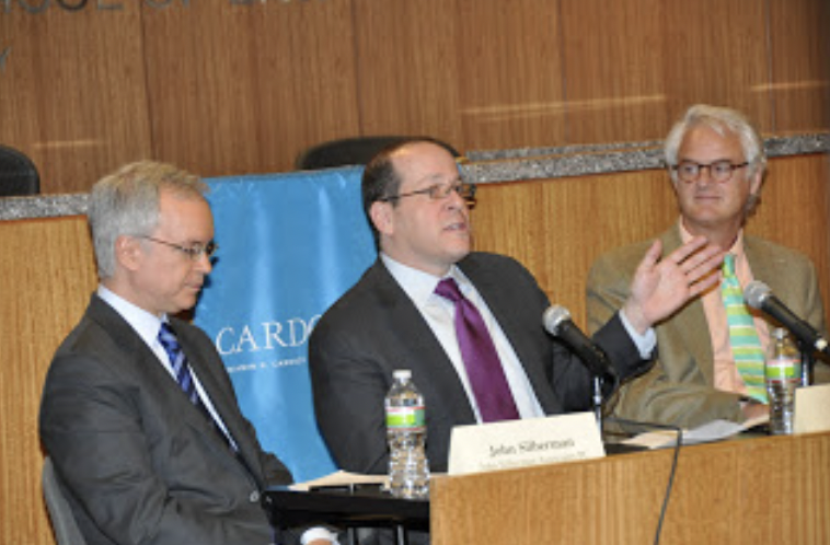Three men in business attire and glasses seated and speaking at a panel.